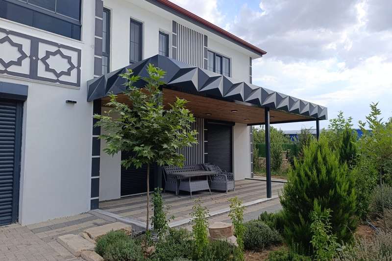 Villanın bahçe cephesine yaslanan antrasit veranda; alınlıkta üç boyutlu kırılımlı alüminyum kompozit panel, tavanda ahşap desenli kaplama ve gömme spotlar, altında rattan oturma grubu
